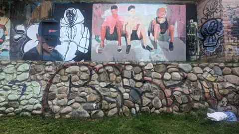 Painted portrait figures and graffiti cover a stone wall.