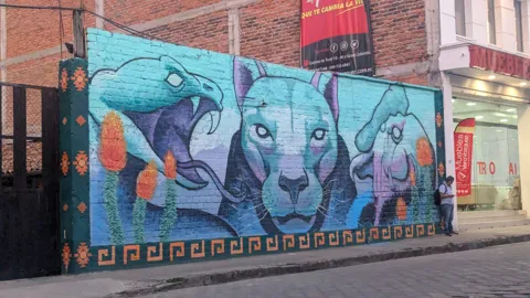 A blue mural with stylized figures covers a street wall.