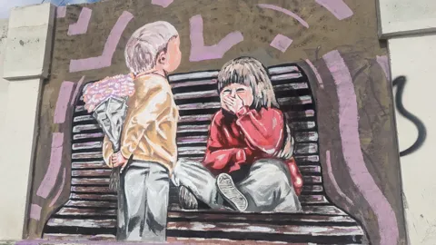 A wall mural shows two seated figures and stylized steps.