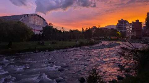 A river is framed by trees and buildings under a colorful sunset.