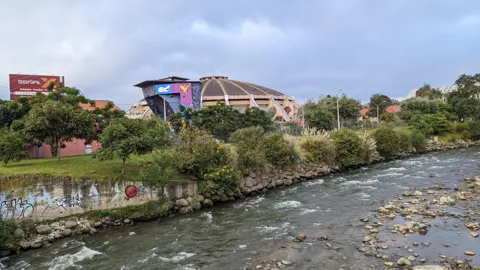 A river flows past city buildings and a low bridge under a cloudy sky.