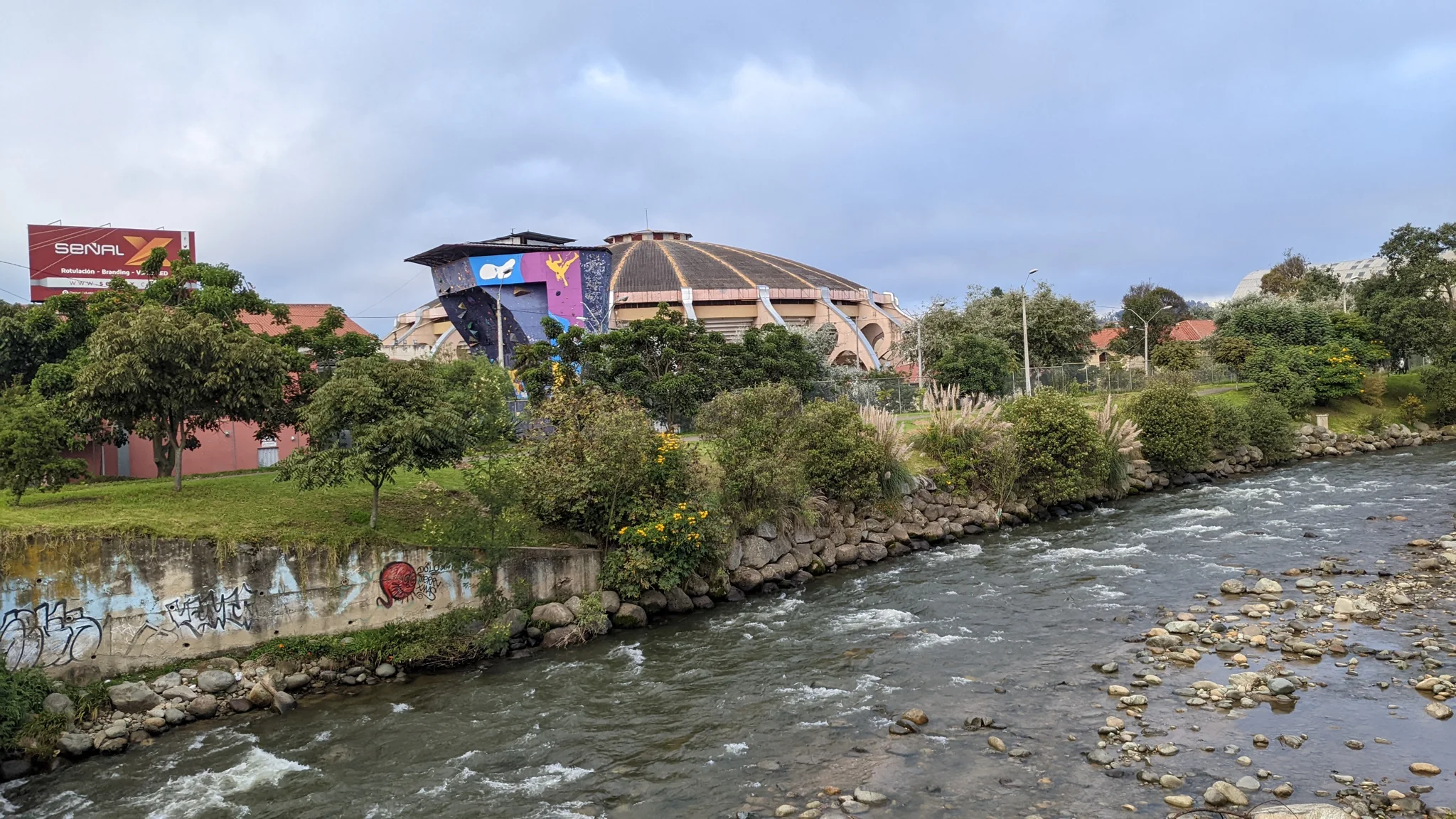 A river flows past city buildings and a low bridge under a cloudy sky.