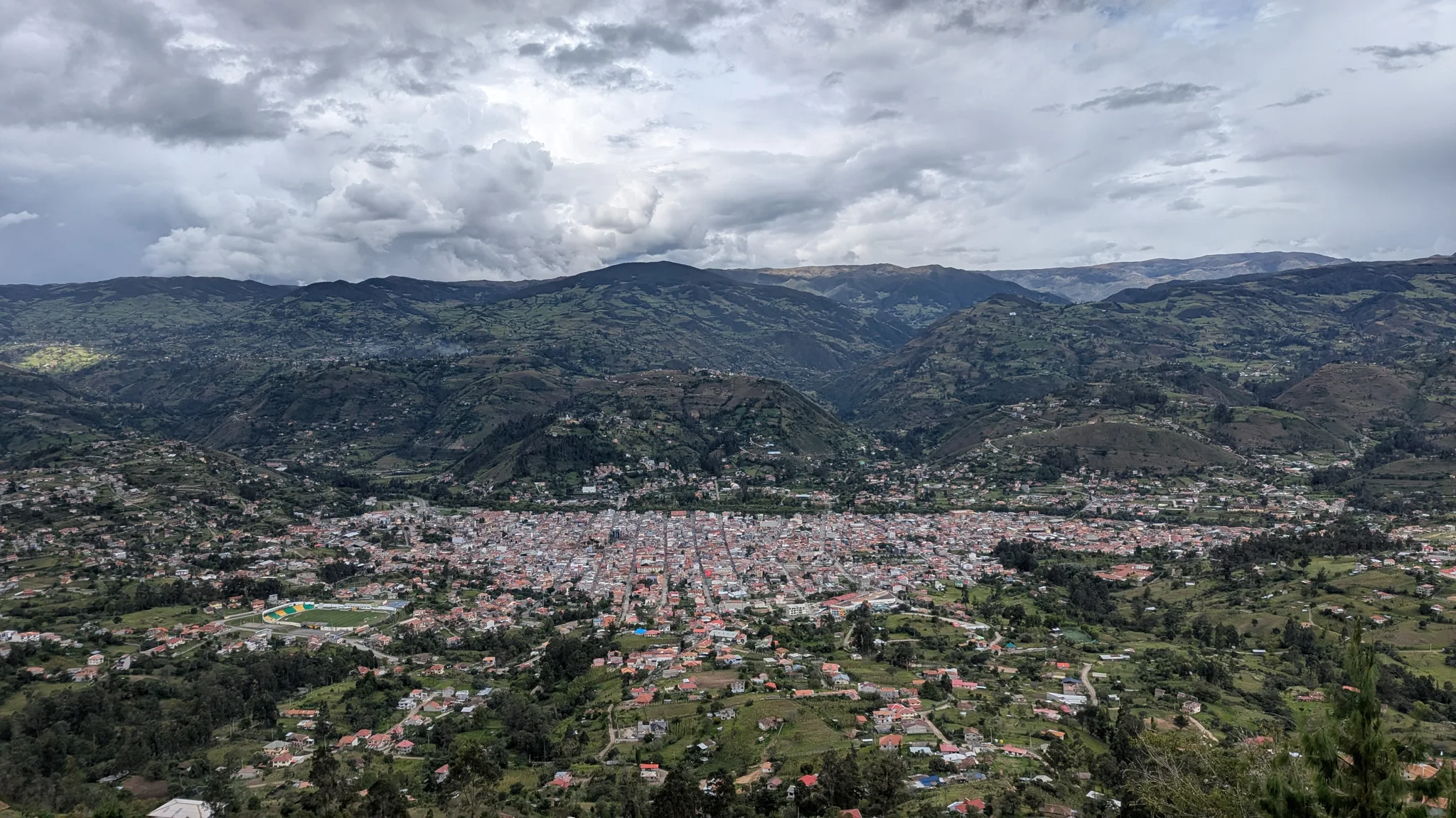 A city valley stretches below rolling green hills and cloudy sky.