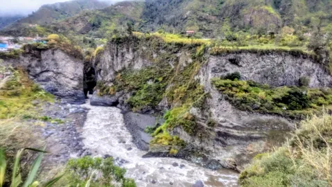 Water cascades over layered rocks through a green highland valley.