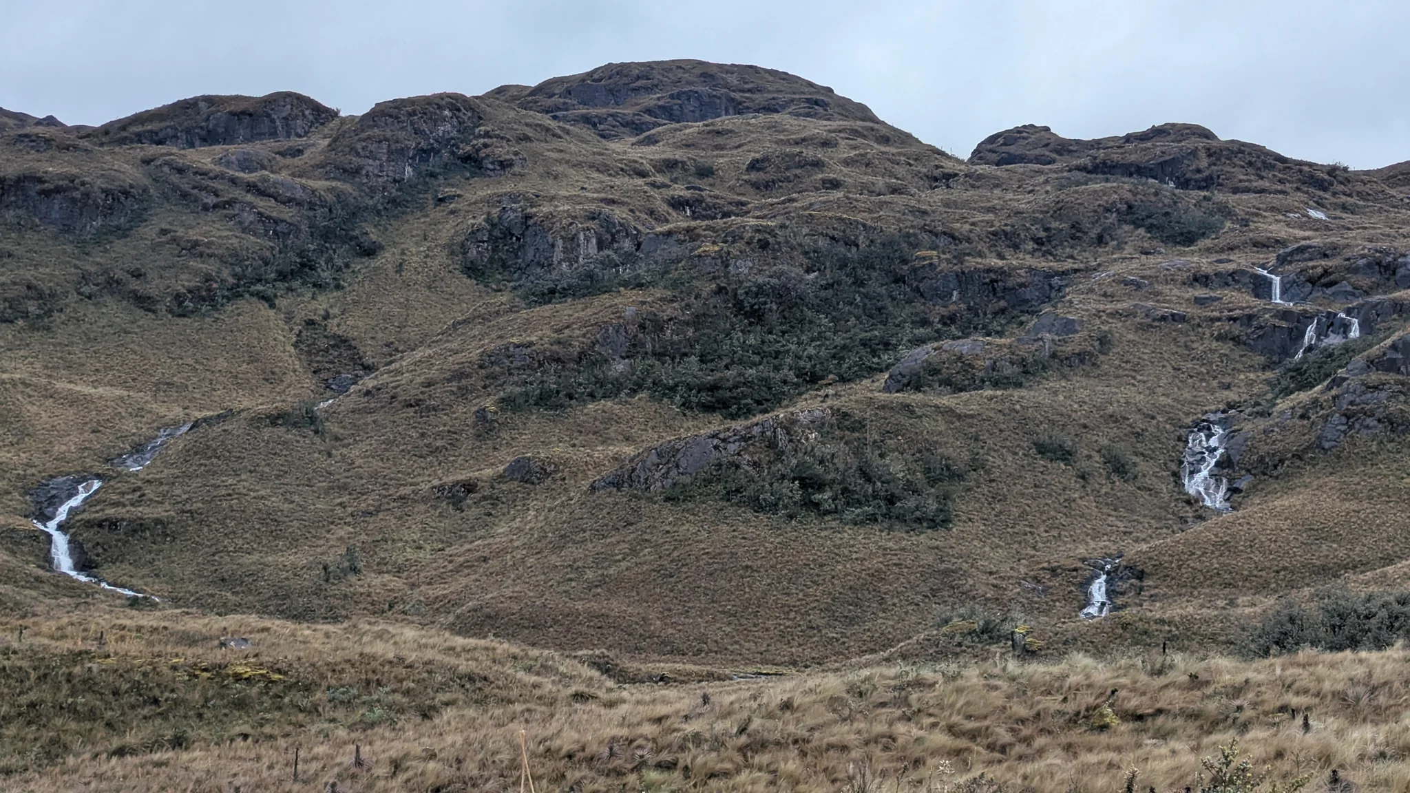 Two separate ribbons of white water descend a broad hillside between páramo grass and rocky shelves.