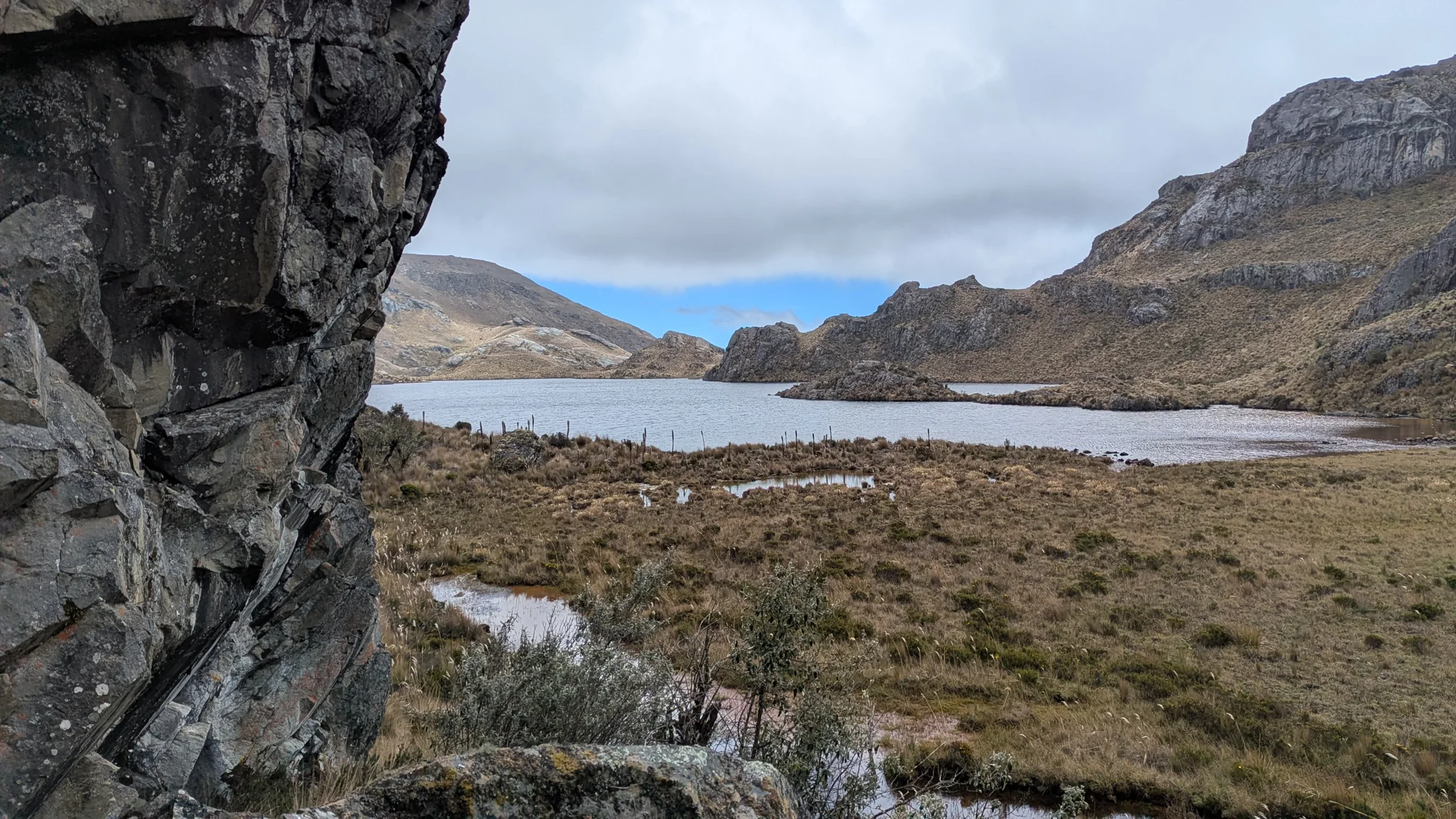 A dark foreground wall frames Estrellascocha de Quitahuaycu and the open grassland along its shore.