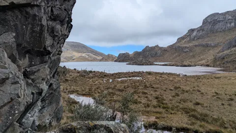A dark foreground wall frames Estrellascocha de Quitahuaycu and the open grassland along its shore.