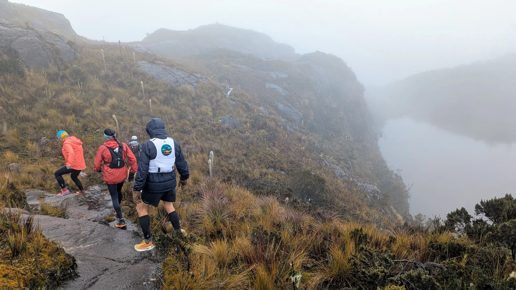 Hikers descend a rocky path beside still water as mist hides the far shore.