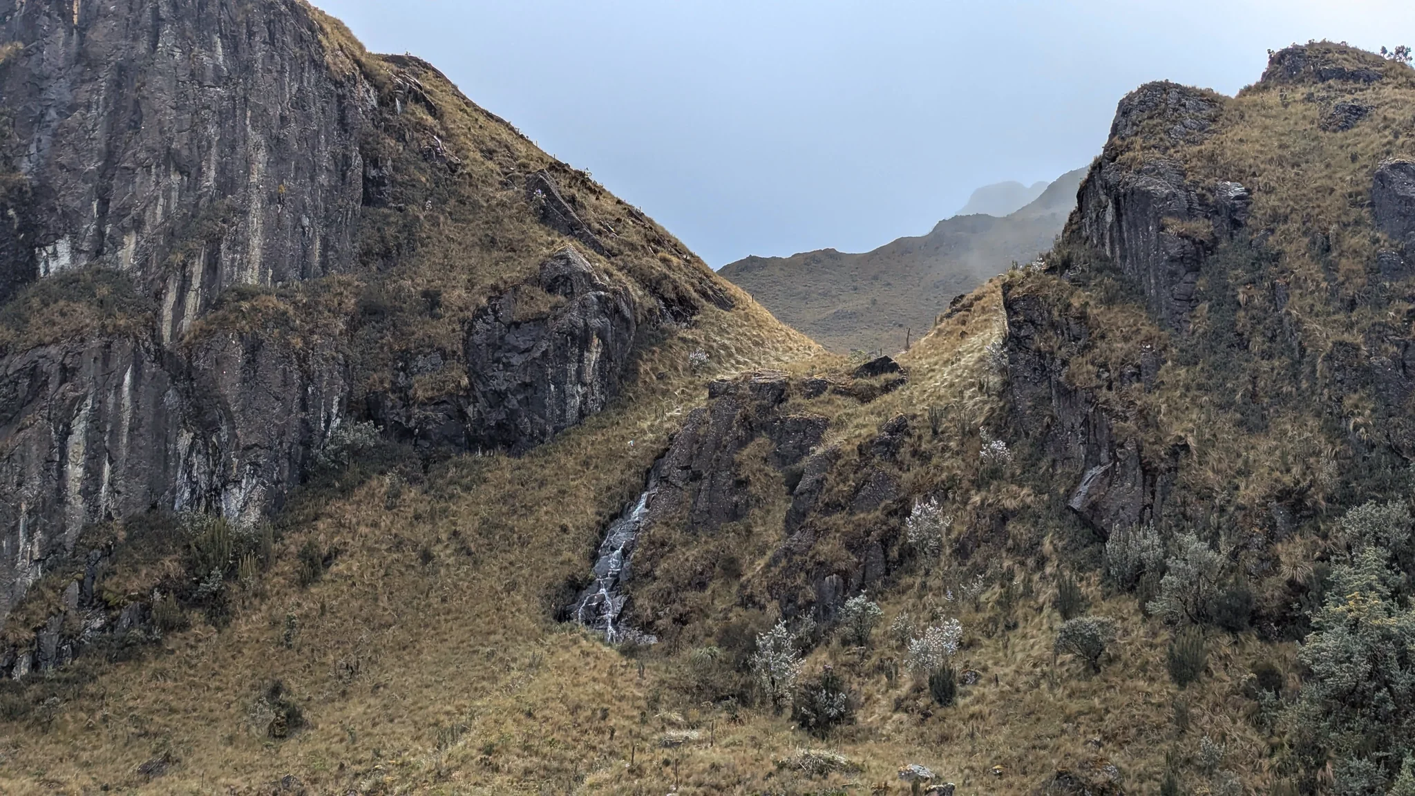 A thin white cascade descends through a cleft between steep dark rock faces and grass-covered slopes.